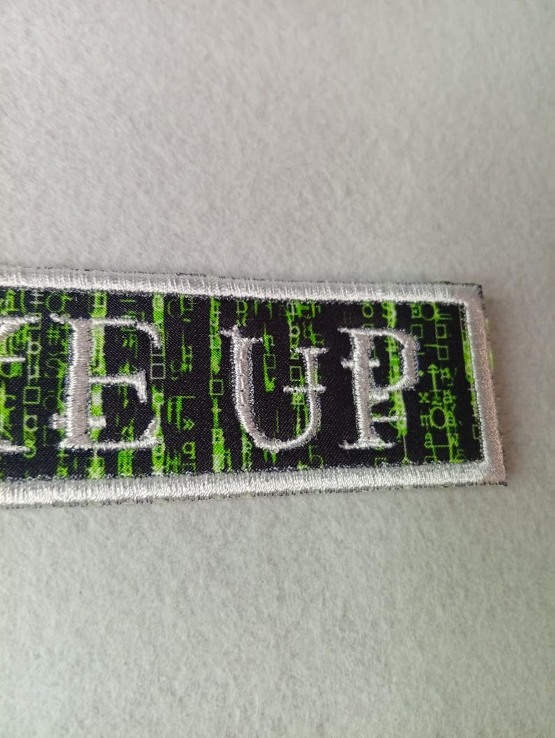 Pode incluir: Patch retangular com a palavra "GEE UP" bordada em fio prateado. O fundo &eacute; preto com um padr&atilde;o de chuva digital verde. O patch tem uma borda branca.