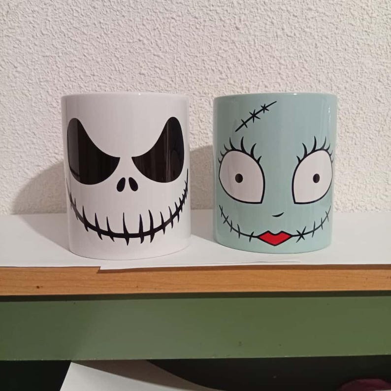 May include: Two ceramic mugs featuring character designs. One mug is white with a black Jack Skellington face. The other is light blue with a Sally face design. Both mugs are cylindrical and sit on a wooden shelf.
