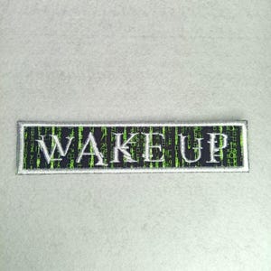 Pode incluir: Um patch retangular com a palavra "WAKE UP" bordada em letras mai&uacute;sculas brancas. O fundo &eacute; preto com padr&otilde;es de c&oacute;digo digital verde. O patch tem uma borda prateada e est&aacute; sobre um fundo cinza claro.