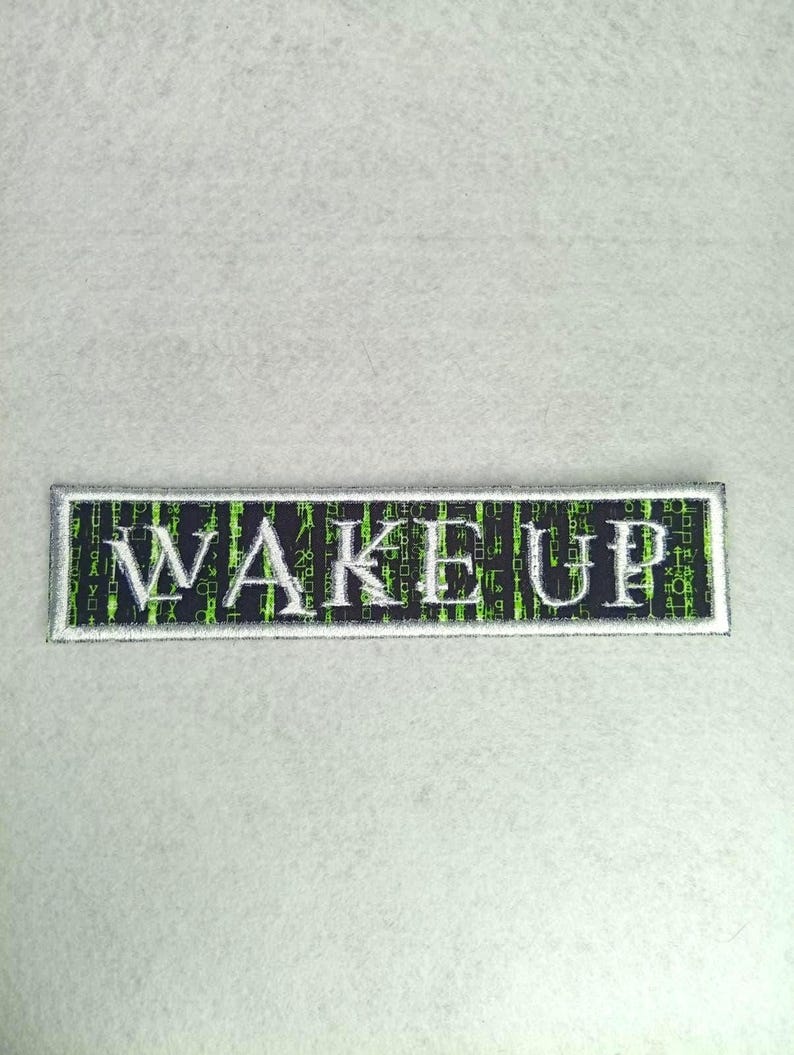 Pode incluir: Um patch retangular com a palavra "WAKE UP" bordada em letras mai&uacute;sculas brancas. O fundo &eacute; preto com padr&otilde;es de c&oacute;digo digital verde. O patch tem uma borda prateada e est&aacute; sobre um fundo cinza claro.