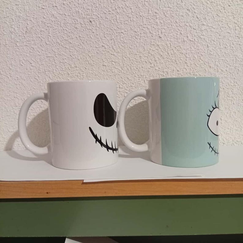 May include: Two white ceramic mugs with black and teal character designs. One mug features a black oval eye and a stitched smile. The other mug has a teal face with a stitched eye and smile. Both mugs have white handles.