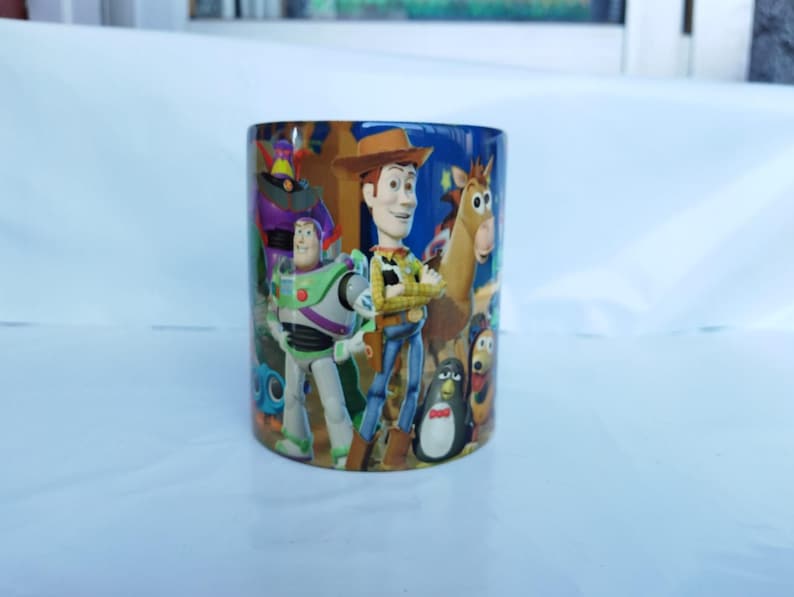 May include: A cylindrical ceramic mug featuring characters from the animated film "Toy Story." The mug showcases Woody, Buzz Lightyear, and other characters in vibrant colors. The mug is suitable for hot or cold beverages.