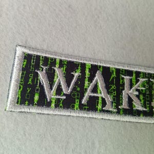 Pode incluir: Patch bordado retangular com a palavra "WAKE" em fio prateado. O fundo &eacute; preto com um padr&atilde;o de c&oacute;digo bin&aacute;rio verde. Possui uma borda prateada.