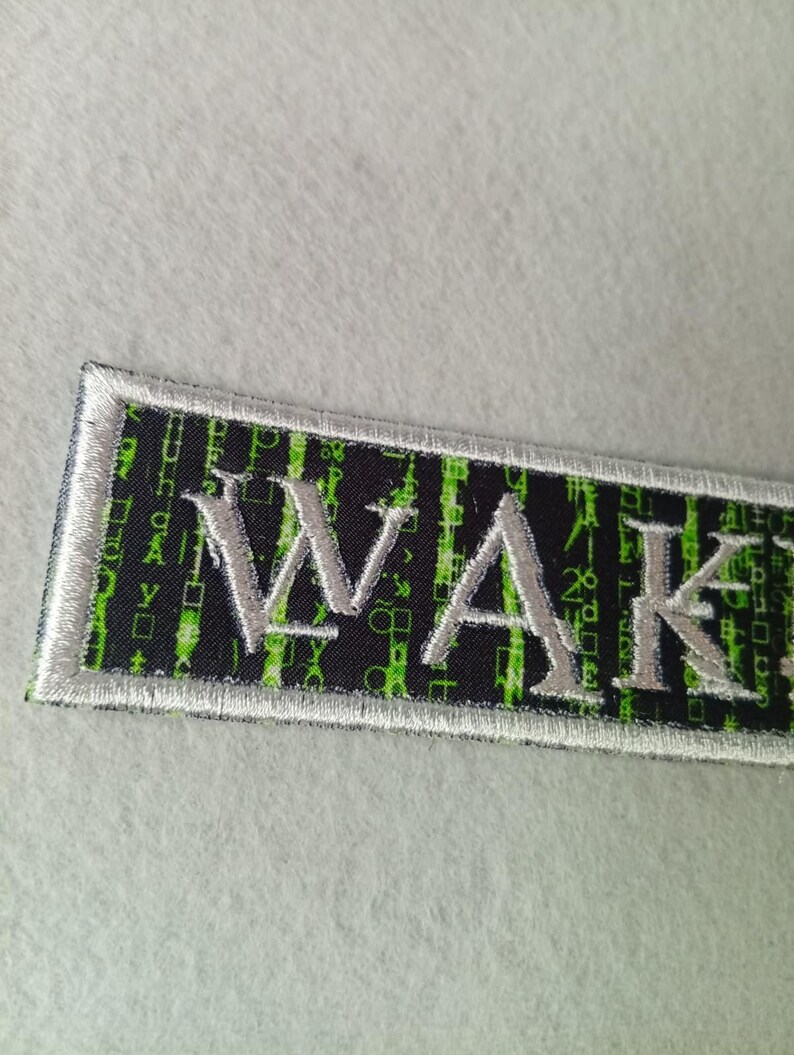 Pode incluir: Patch bordado retangular com a palavra "WAKE" em fio prateado. O fundo &eacute; preto com um padr&atilde;o de c&oacute;digo bin&aacute;rio verde. Possui uma borda prateada.