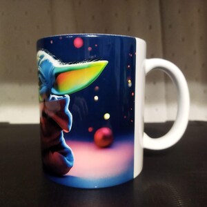 K&ouml;nnte beinhalten: Wei&szlig;er Keramikbecher mit einer farbenfrohen Illustration einer Figur mit gro&szlig;en Ohren und blauem Gesicht vor einem dunkelblauen Hintergrund mit roten und gelben Kugeln. Der Becher hat einen wei&szlig;en Henkel.