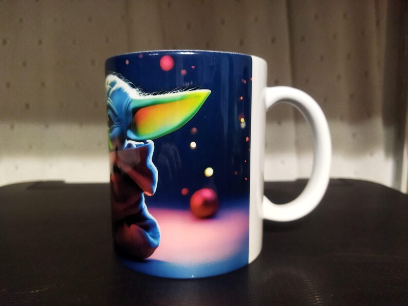 K&ouml;nnte beinhalten: Wei&szlig;er Keramikbecher mit einer farbenfrohen Illustration einer Figur mit gro&szlig;en Ohren und blauem Gesicht vor einem dunkelblauen Hintergrund mit roten und gelben Kugeln. Der Becher hat einen wei&szlig;en Henkel.