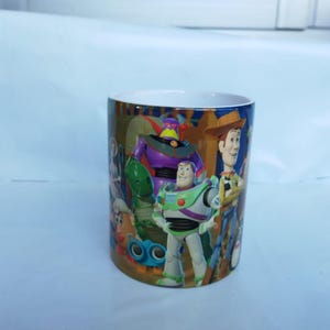 May include: A cylindrical ceramic mug featuring characters from the animated film "Toy Story." The mug showcases Buzz Lightyear, Woody, and other characters in vibrant colors. The mug has a white interior and a glossy finish.