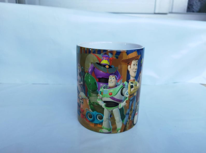 May include: A cylindrical ceramic mug featuring characters from the animated film "Toy Story." The mug showcases Buzz Lightyear, Woody, and other characters in vibrant colors. The mug has a white interior and a glossy finish.