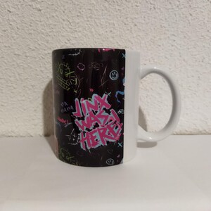 May include: A black and white coffee mug with a colorful graphic design. The design features the text "Jinx was here" in pink letters with other colorful graffiti-style markings.