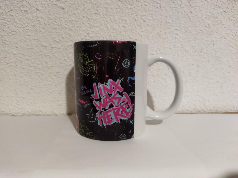 May include: A black and white coffee mug with a colorful graphic design. The design features the text "Jinx was here" in pink letters with other colorful graffiti-style markings.