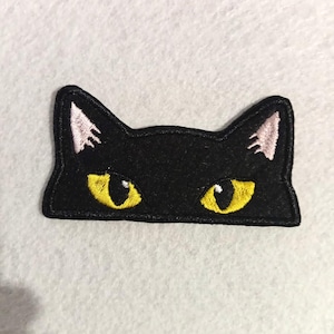 Può includere: Toppa a forma di gatto nero ricamata con occhi gialli e interno orecchie rosa. La toppa ha la forma della metà superiore del muso di un gatto. Ideale per personalizzare abbigliamento o accessori.