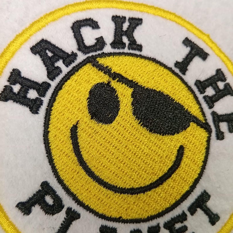 May include: Embroidered patch featuring a yellow smiley face wearing black sunglasses. The phrase "HACK THE PLANET" is embroidered in black around the smiley face on a white background with a yellow border.