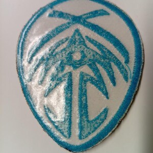 Peut inclure: &Eacute;cusson brod&eacute; bleu et blanc avec un motif stylis&eacute;. Le patch a un fond blanc avec une bordure bleue et un motif central compos&eacute; de formes et de lignes g&eacute;om&eacute;triques.