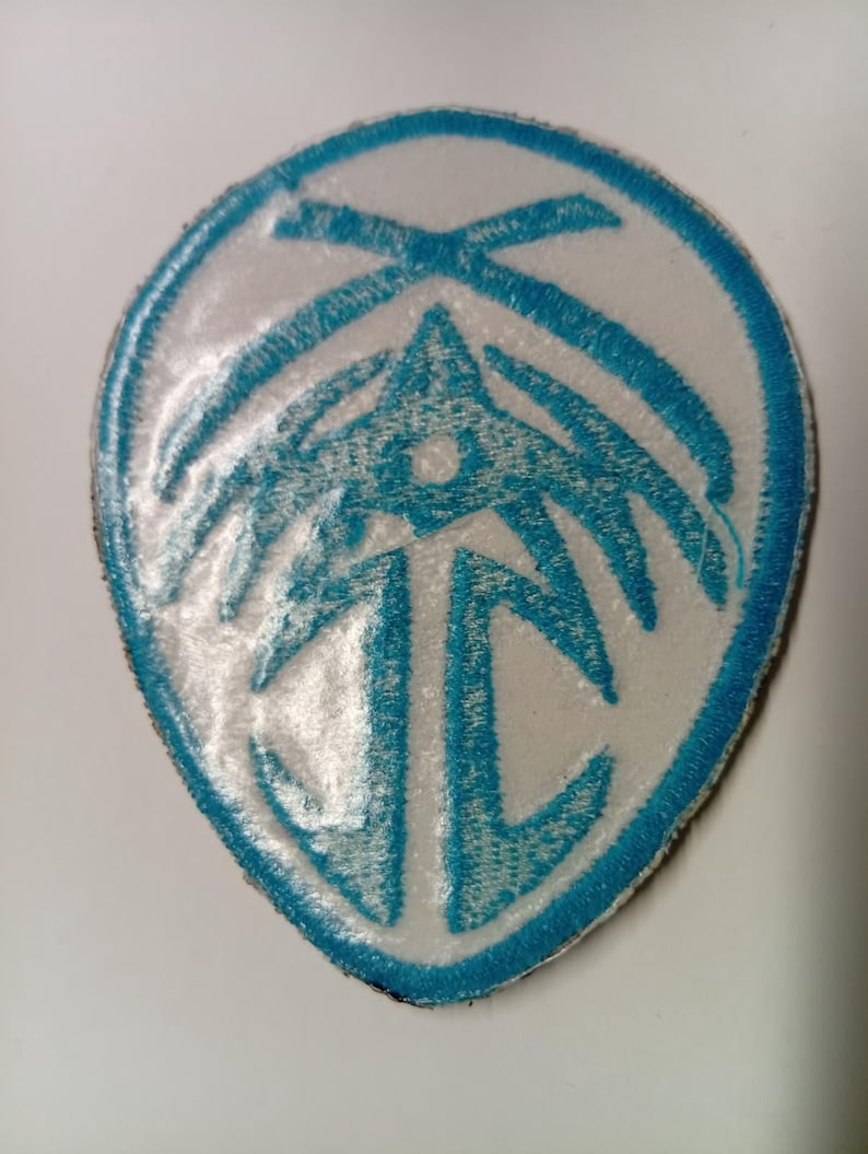 Peut inclure: &Eacute;cusson brod&eacute; bleu et blanc avec un motif stylis&eacute;. Le patch a un fond blanc avec une bordure bleue et un motif central compos&eacute; de formes et de lignes g&eacute;om&eacute;triques.