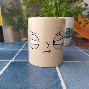 Peut inclure: Une tasse en céramique beige avec un visage de dessin animé dessiné dessus. Le visage a deux yeux avec des pupilles noires et des sourcils, et une bouche légèrement froncée.