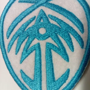 Peut inclure: &Eacute;cusson brod&eacute; avec un motif turquoise sur fond blanc. Le motif turquoise pr&eacute;sente un embl&egrave;me stylis&eacute; avec une forme de fl&egrave;che centrale et une bordure ovale. L'&eacute;cusson est probablement destin&eacute; aux v&ecirc;tements ou aux accessoires.