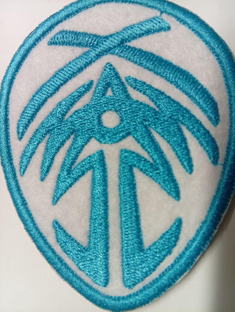 Peut inclure: &Eacute;cusson brod&eacute; avec un motif turquoise sur fond blanc. Le motif turquoise pr&eacute;sente un embl&egrave;me stylis&eacute; avec une forme de fl&egrave;che centrale et une bordure ovale. L'&eacute;cusson est probablement destin&eacute; aux v&ecirc;tements ou aux accessoires.