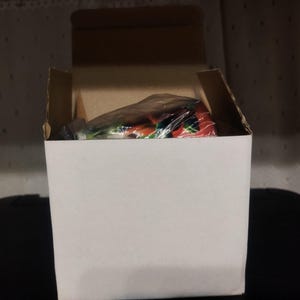 K&ouml;nnte beinhalten: Ein wei&szlig;er Karton mit ge&ouml;ffneten oberen Klappen, der einen verpackten Artikel im Inneren zeigt. Der Artikel ist teilweise sichtbar und zeigt rote, gr&uuml;ne und wei&szlig;e Farben. Die Box steht auf einer schwarzen Oberfl&auml;che.