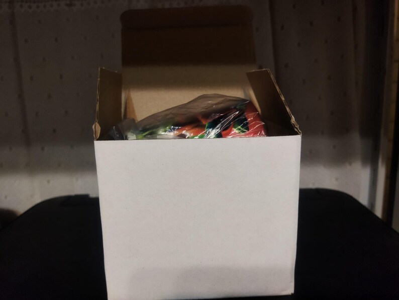 K&ouml;nnte beinhalten: Ein wei&szlig;er Karton mit ge&ouml;ffneten oberen Klappen, der einen verpackten Artikel im Inneren zeigt. Der Artikel ist teilweise sichtbar und zeigt rote, gr&uuml;ne und wei&szlig;e Farben. Die Box steht auf einer schwarzen Oberfl&auml;che.