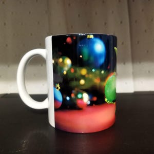 K&ouml;nnte beinhalten: Wei&szlig;e Keramiktasse mit einem festlichen Weihnachtsbaumschmuck-Design. Die Tasse zeigt bunte Ornamente in Blau, Gr&uuml;n und Rot vor einem dunklen Hintergrund mit Bokeh-Lichtern. Der Griff ist wei&szlig;.