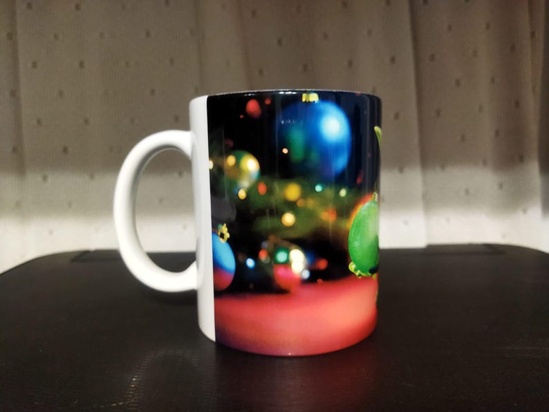 K&ouml;nnte beinhalten: Wei&szlig;e Keramiktasse mit einem festlichen Weihnachtsbaumschmuck-Design. Die Tasse zeigt bunte Ornamente in Blau, Gr&uuml;n und Rot vor einem dunklen Hintergrund mit Bokeh-Lichtern. Der Griff ist wei&szlig;.