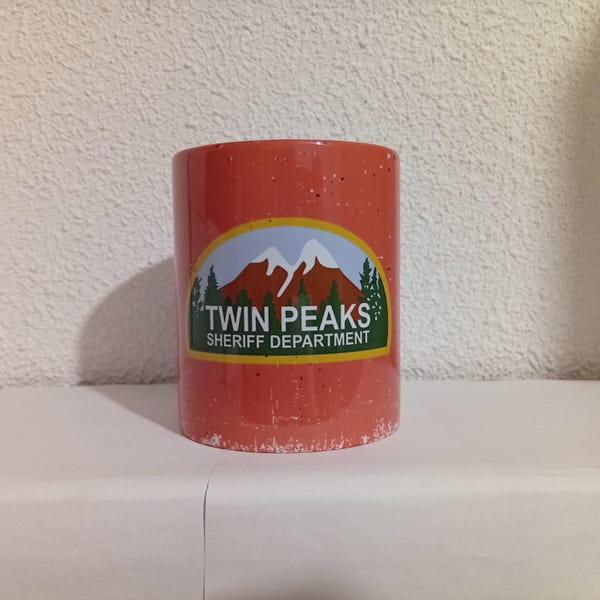 Puede incluir: Una taza cilíndrica roja moteada con el logotipo "Twin Peaks Sheriff Department". El logotipo incluye una cordillera, árboles y el texto "Twin Peaks" y "Sheriff Department" en blanco y verde.