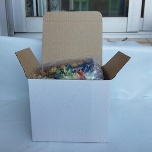 May include: A white cardboard box with a brown cardboard lid and flaps. Inside, a clear plastic bag contains small, colorful figurines. The box is open, revealing the contents, and sits against a white surface.
