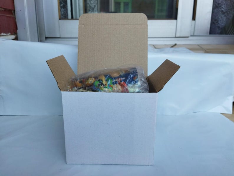 May include: A white cardboard box with a brown cardboard lid and flaps. Inside, a clear plastic bag contains small, colorful figurines. The box is open, revealing the contents, and sits against a white surface.