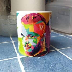 Könnte beinhalten: Eine weiße Keramiktasse mit einem farbenfrohen Pop-Art-Design eines Mannes mit Bart und roten Haaren. Das Design umfasst eine Vielzahl von Farben, darunter Rot, Gelb, Blau, Grün und Lila.