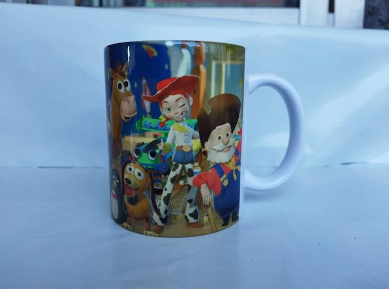 May include: White ceramic mug featuring characters from the animated film Toy Story. The mug has a white handle and is decorated with images of Woody, Jessie, Buzz Lightyear, and other characters from the movie.