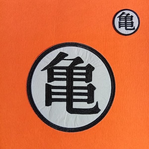 Puede incluir: Dos parches bordados circulares sobre un fondo de tela naranja brillante. Cada parche presenta una base circular blanca con un borde negro grueso y el carácter kanji japonés negro para tortuga, también conocido como el símbolo Kame.
