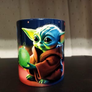 K&ouml;nnte beinhalten: Keramikbecher mit einer lebendigen Illustration einer Figur aus einem beliebten Science-Fiction-Franchise. Die Figur mit gr&uuml;ner Haut und gro&szlig;en Augen h&auml;lt eine gr&uuml;ne Kugel. Der Becher hat einen dunkelblauen Hintergrund mit roter Basis.