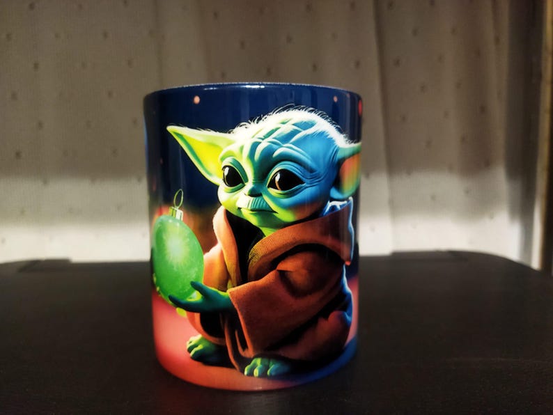 K&ouml;nnte beinhalten: Keramikbecher mit einer lebendigen Illustration einer Figur aus einem beliebten Science-Fiction-Franchise. Die Figur mit gr&uuml;ner Haut und gro&szlig;en Augen h&auml;lt eine gr&uuml;ne Kugel. Der Becher hat einen dunkelblauen Hintergrund mit roter Basis.