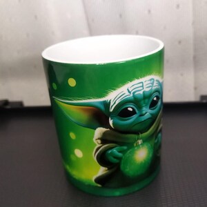 Pu&ograve; includere: Tazza in ceramica verde con un'illustrazione dettagliata di un personaggio Grogu che tiene un ornamento verde brillante. La tazza ha un interno bianco e un esterno verde con dettagli a pois gialli.