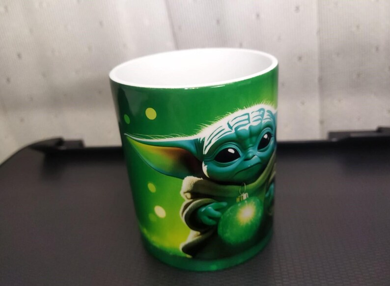 Pu&ograve; includere: Tazza in ceramica verde con un'illustrazione dettagliata di un personaggio Grogu che tiene un ornamento verde brillante. La tazza ha un interno bianco e un esterno verde con dettagli a pois gialli.