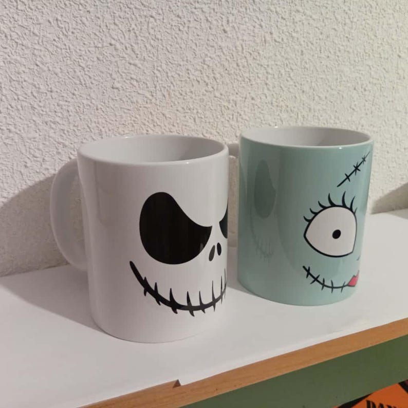 May include: Two ceramic mugs with character faces. One is white with a black skull face, and the other is light blue with a stitched face design. Both mugs have a white interior and a curved handle.