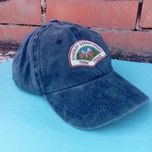Op de afbeelding: Donkerblauwe denim baseballcap met een gebogen klep. De pet heeft een patch met de tekst "SHERIFF DEPARTMENT TWIN PEAKS" en een afbeelding van bergen. De hoed ligt op een lichtblauwe ondergrond.