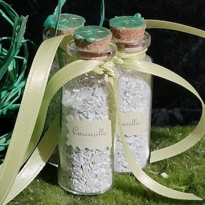 May include: Three small glass bottles with cork stoppers and green clover-shaped lids. The bottles are filled with white, flaky material and tied with a yellow ribbon. The labels on the bottles read "Cascarilla" and "arilla".