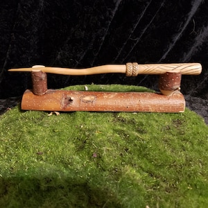 Peut inclure: Une baguette en bois avec un motif sculpt&eacute; sur la poign&eacute;e, pr&eacute;sent&eacute;e sur un support en bois. La baguette est brune et l&eacute;g&egrave;rement incurv&eacute;e. Le support est en bois naturel avec de l'&eacute;corce encore attach&eacute;e.