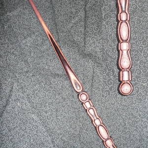 May include: Two wooden wands with intricate carvings and a dark brown and pink color scheme. The wands are lying on a dark gray fabric with a swirling pattern.