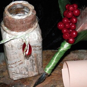 May include: A vintage-style glass jar with a burlap lid and a decorative red bead hanging from the lid. The jar is decorated with white paper and text. A green and white quill pen with a red berry bouquet is next to the jar.