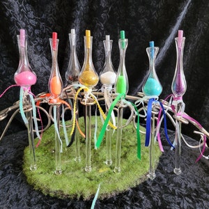 May include: A set of eight glass bottles with colorful glitter inside, each bottle is topped with a stopper and attached to a long, thin, clear wand with a ribbon. The wands are arranged in a row on a green surface.