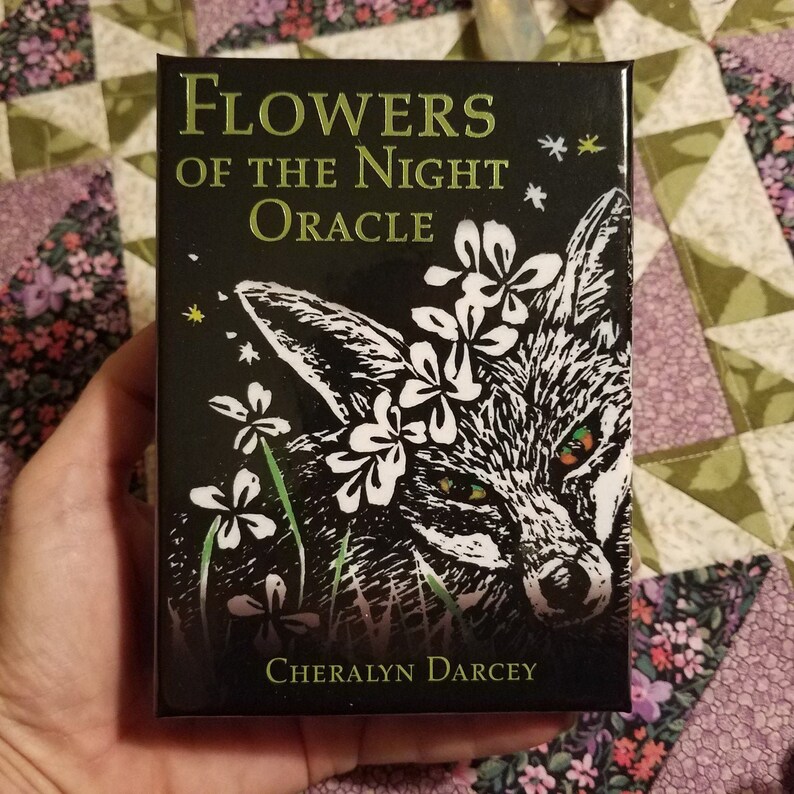 May include: A black box with gold lettering that reads "Flowers of the Night Oracle" and a black and white illustration of a fox with flowers on its head. The box is by Cheralyn Darcey.