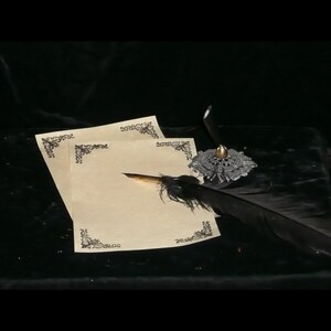 May include: A black feather quill pen rests on a sheet of cream-colored stationery with decorative borders. The pen is placed on a silver inkwell with a gold top.