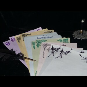May include: A stack of blank stationery paper with decorative borders in black, green, and purple. The paper is arranged in a fan shape with a quill pen on top.