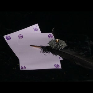 May include: A black feather quill pen with a silver and gold inkwell sits on a purple stationery set with a swirl design.