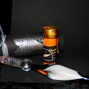 May include: A small orange bottle with a gold cap and a black label that reads "Ghost Ink Invisible Handwriting Ink The Original Gilded Ink". The bottle is sitting on a black surface next to a glass dip pen and a black and silver tube.
