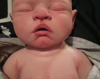 May include: A lifelike, bald baby doll with closed eyes and a slightly open mouth. The doll has a realistic skin tone with subtle shading and details. The doll's chest and shoulders are visible, suggesting a reborn doll.