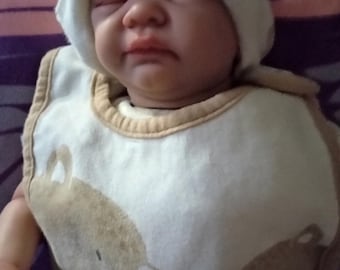 Puede incluir: Un bebé con un gorro blanco con un pequeño parche y un babero con un diseño de oso. El babero es blanco con una ilustración de oso marrón y un ribete beige. Los ojos del bebé están cerrados.