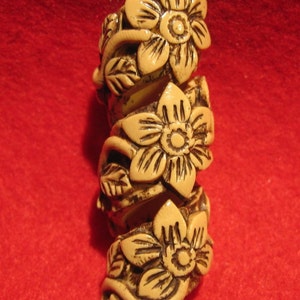 May include: A beige, floral-themed decorative object. The object is a spiral shape with three prominent flowers and leaves. The flowers have detailed petals and are accented with dark brown lines. The background is a solid red.